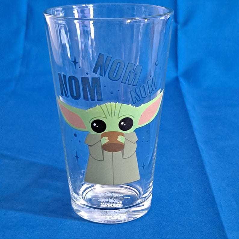 Peut inclure: Verre &agrave; bi&egrave;re transparent avec une image de dessin anim&eacute; de Grogu, &eacute;galement connu sous le nom de Baby Yoda, tenant une tasse. Les mots "NOM NOM" sont imprim&eacute;s en bleu au-dessus du personnage. Le verre a un logo Star Wars en bas.