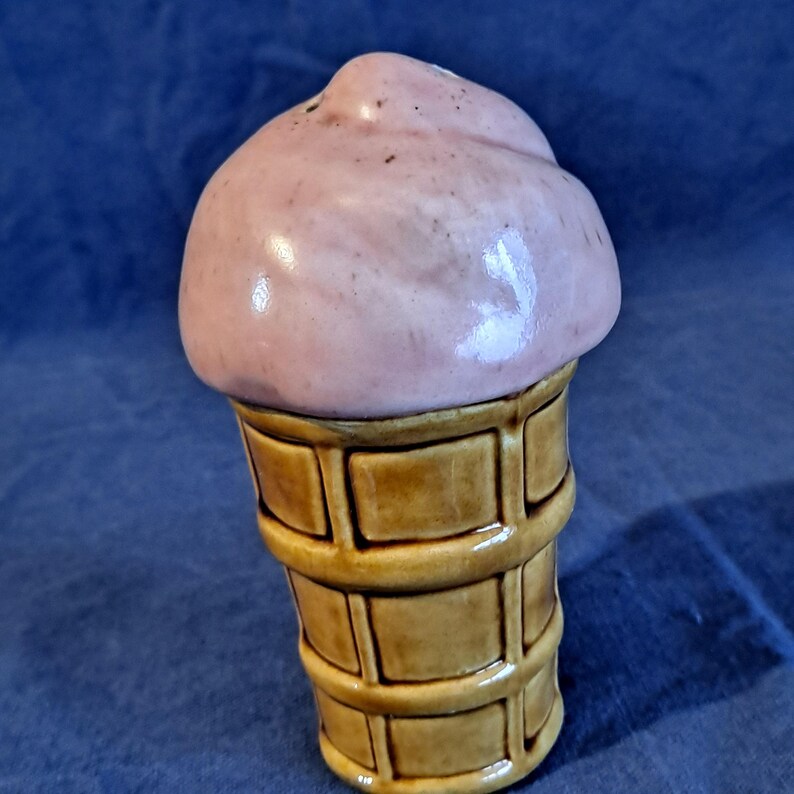 K&ouml;nnte beinhalten: Ein Keramik-Salz- und Pfefferstreuer-Set in Form einer Eist&uuml;te. Die Eiskugel ist hellrosa und die Waffel ist hellbraun mit einem Waffelmuster. Das Set ist so gestaltet, dass es wie eine Kugel Eis auf einer Waffel aussieht.