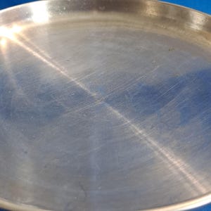 May include: A round, silver-colored metal plate with a slightly raised edge. The plate has a brushed texture with visible scratches and marks across its surface. The plate is set against a blue background.