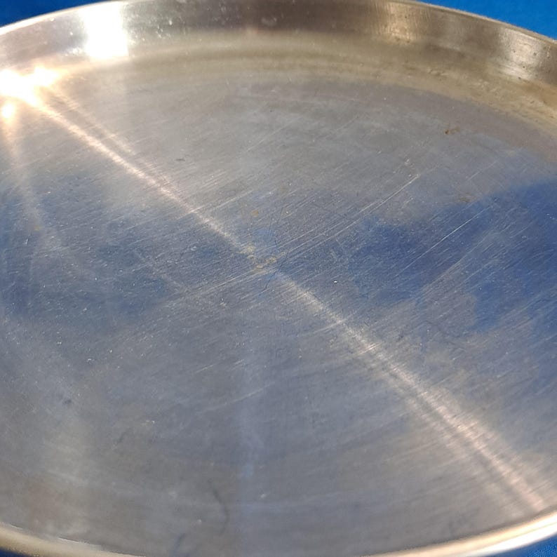May include: A round, silver-colored metal plate with a slightly raised edge. The plate has a brushed texture with visible scratches and marks across its surface. The plate is set against a blue background.