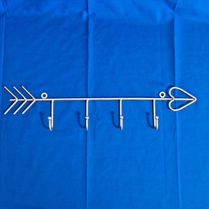 May include: A metal arrow-shaped wall hook with four hooks. The arrow has a heart-shaped tip and a series of lines forming the fletching. The hooks are evenly spaced along the shaft. The metal is a light silver color.