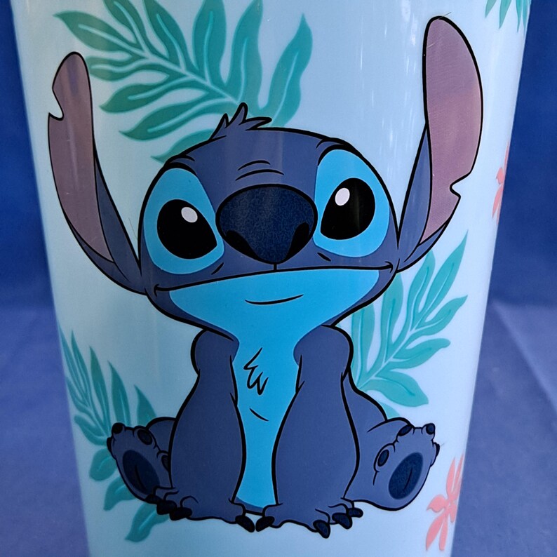 May include: A light blue tumbler featuring a cartoon image of Stitch from Lilo & Stitch. The character is blue with large black eyes and large ears. The tumbler has a floral and leaf pattern in light green and pink.