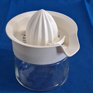 May include: A white plastic citrus juicer with a ribbed reamer and a built-in strainer sits atop a clear glass jar. The juicer has a pouring spout and is designed for easy juice extraction. The product is designed for kitchen use.