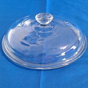 Könnte beinhalten: Ein klarer Glasdeckel mit einem kleinen, runden Griff. Der Deckel ist kreisförmig mit einer leicht gewölbten Form und einer Reihe konzentrischer Ringe in der Nähe der Mitte. Der Griff ist ebenfalls aus klarem Glas.