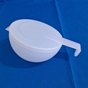 Puede incluir: Tazón de plástico blanco con tapa y asa. La tapa tiene la palabra "Microwave" impresa.