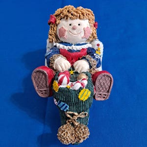Puede incluir: Figura de muñeca de cerámica con una cara sonriente, vestida con un vestido azul y rojo, y sosteniendo una cesta verde llena de caramelos. La muñeca tiene cabello castaño, lazos rojos y mejillas rosadas. La figura está sentada en una silla blanca.