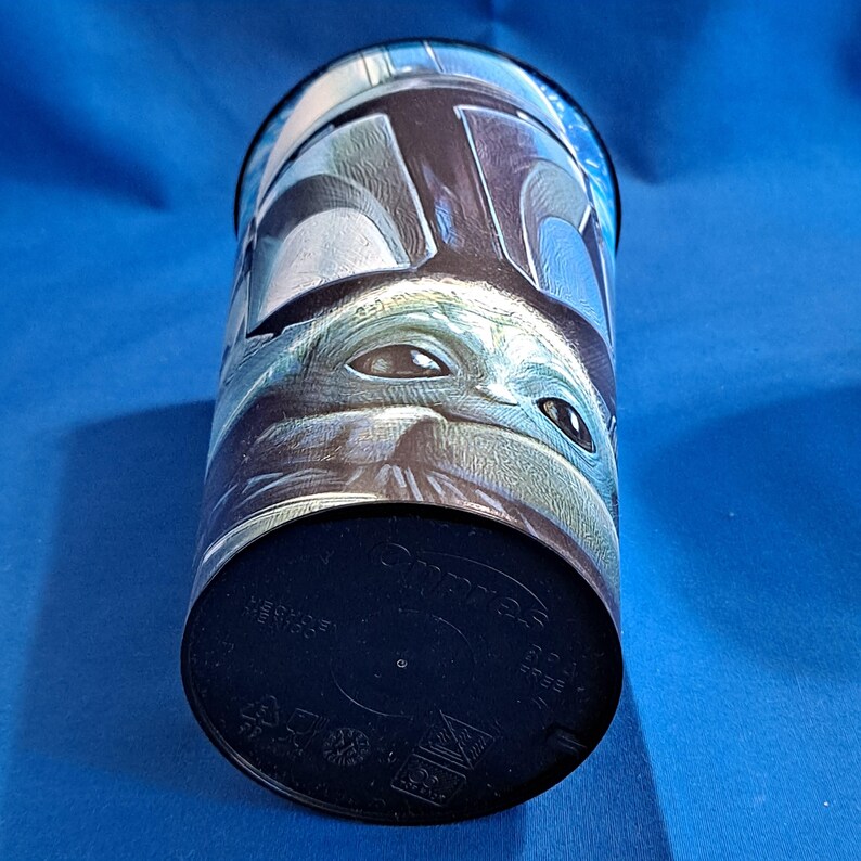 May include: A cylindrical tumbler featuring a detailed image of The Mandalorian and Grogu from the Star Wars series. The design wraps around the cup, showcasing the characters' faces in shades of blue, gray, and green. The bottom is black.
