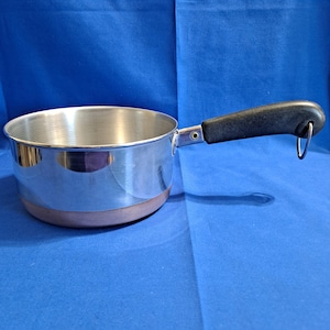 May include: A small, shiny, stainless steel saucepan with a copper-colored base and a black handle. The handle has a loop for hanging. The pan is sitting on a blue surface.