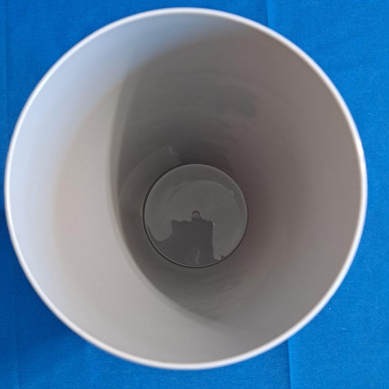 May include: An overhead view of a light grey cylindrical container with a dark grey interior. The container has a wide opening and a flat base. The background is a solid blue colour.