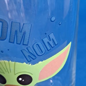 Peut inclure: Verre transparent avec un fond bleu, orn&eacute; d'un dessin de personnage vert et rose. Les mots "NOM NOM" sont imprim&eacute;s en bleu. Le dessin comprend une oreille et un &oelig;il de personnage de dessin anim&eacute;.