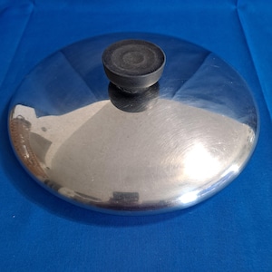 May include: A stainless steel pot lid with a black, circular handle. The lid is round and reflective, with a brushed metal finish. The handle is made of a dark, possibly rubbery material.