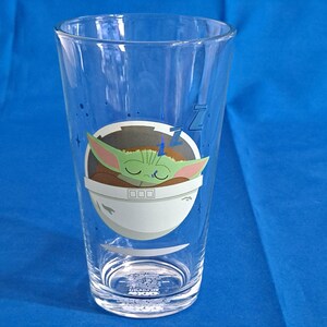 Peut inclure: Un verre transparent avec une image de dessin anim&eacute; d'un personnage Grogu endormi dans un pod blanc et gris. Le verre a une forme haute et &eacute;lanc&eacute;e. Le fond est bleu.