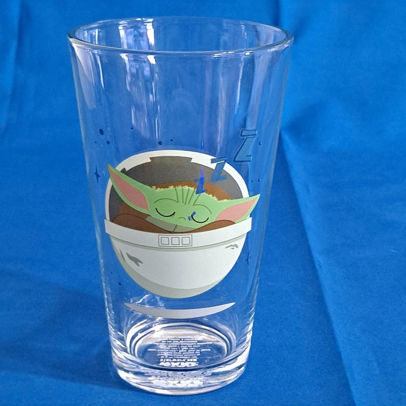 Peut inclure: Un verre transparent avec une image de dessin anim&eacute; d'un personnage Grogu endormi dans un pod blanc et gris. Le verre a une forme haute et &eacute;lanc&eacute;e. Le fond est bleu.
