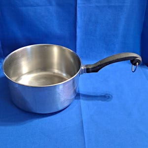 Könnte beinhalten: Ein Edelstahltopf mit schwarzem Griff und einer silberfarbenen Metallschlaufe. Der Topf ist leer und steht auf einer blauen Oberfläche. Der Griff ist gebogen und an der Seite des Topfes befestigt.