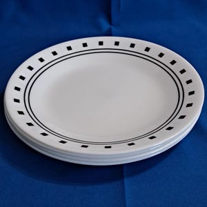May include: A stack of white oval plates with a black border and small black square accents. The plates are stacked on top of each other. The background is a solid blue. The plates are approximately 25 cm in diameter.