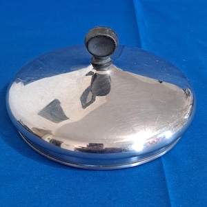 May include: A circular, polished stainless steel lid featuring a smooth, reflective surface and a centered, matte black, spherical knob handle. The lid has a slightly domed shape with a clean, metallic finish, designed to fit securely on cookware.