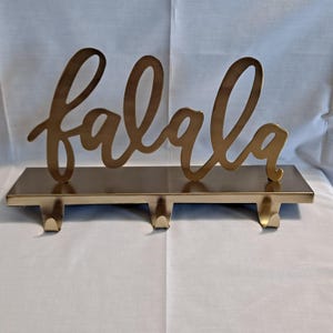 May include: A gold-toned metal wall hook rack featuring the word falala in a cursive script font. The horizontal base plate supports three downward-curving hooks for hanging items, all finished in a smooth, reflective metallic gold surface.