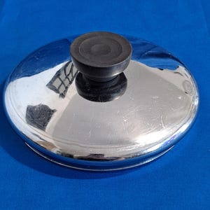May include: A shiny, chrome-colored lid with a black, round handle. The lid reflects the surrounding environment, showing a distorted image. The handle is centered on the lid, which appears to be made of metal.