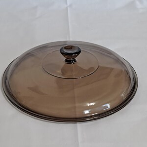 May include: A clear, brown-tinted glass lid with a rounded shape and a small, dark brown knob. The lid is designed to fit a pot or pan, offering a view of the contents while cooking. Kitchenware.