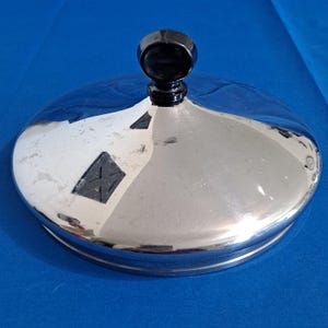 Pode incluir: Tampa prateada brilhante com um botão preto na parte superior. A tampa tem formato circular e uma superfície refletora. O botão é redondo e liso. A tampa parece ser de metal.