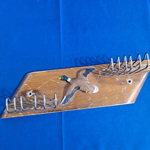 May include: Wooden wall-mounted key holder with a decorative flying duck. The key holder has a light brown wooden base with metal key hooks. The duck is painted with brown, white, green, and yellow details. The key holder is mounted on a blue background.