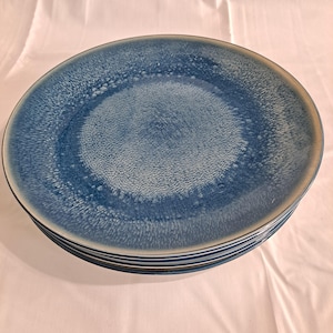 Op de afbeelding: Een stapel blauwe keramische borden met een gespikkelde textuur. De borden hebben een kleurovergang, met een lichter blauw midden en een donkerdere blauwe rand. De borden zijn op elkaar gestapeld.
