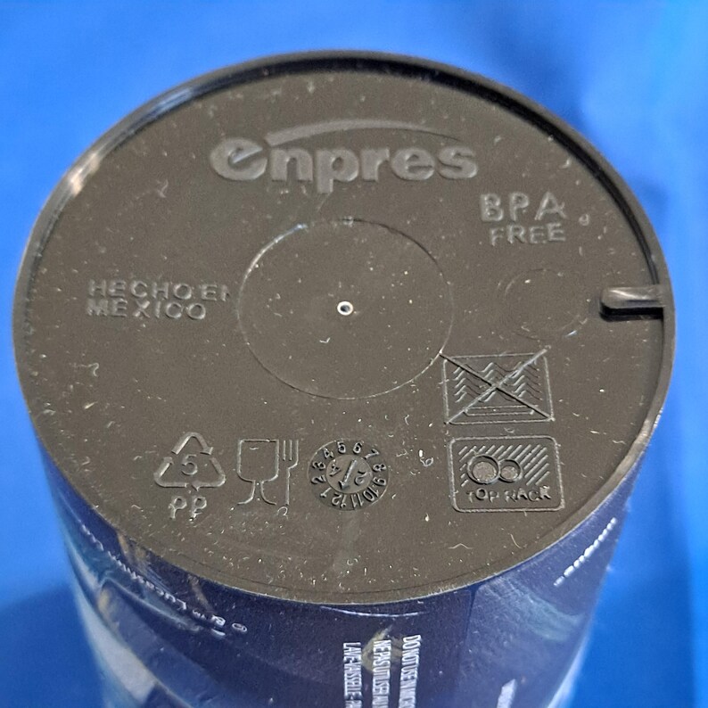 May include: A close-up of a black plastic lid with the embossed text "enpres" and "BPA FREE". The lid also features the text "HECHO EN MEXICO" and several recycling symbols. The lid is for a container.
