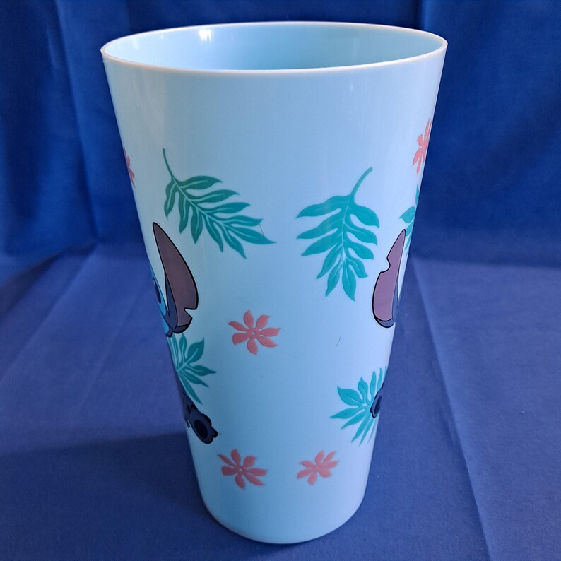 May include: A light blue plastic tumbler featuring a cartoon character design. The tumbler is decorated with images of the character, green leaves, and pink flowers. The tumbler is cylindrical and has a wide opening.