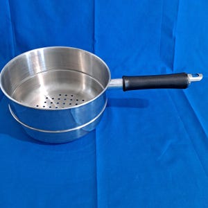 May include: A stainless steel steamer pot with a black handle. The pot has a perforated insert and a handle with a loop for hanging. The pot is designed for steaming vegetables or other foods.