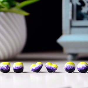 Könnte beinhalten: Sechs kleine Ohrringe in Form von Quadraten und Herzen. Die Ohrringe sind in den Farben der Non-Binary-Pride-Flagge bemalt: Gelb, Weiß, Lila und Schwarz.