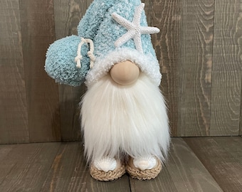 May include: A blue and white gnome with a white starfish on its hat. The gnome is wearing brown sandals with white seashells on them. The gnome is standing on a wooden surface.