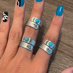 May include: Three silver-toned wrap rings with a turquoise stone and stamped initials. Each ring has a heart symbol and a letter. The rings are displayed on a hand with blue nail polish.