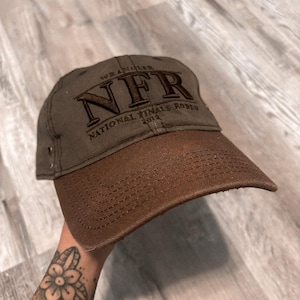 May include: An olive green baseball cap with a brown brim. The front features dark brown embroidered text that reads Wrangler NFR National Finals Rodeo 2012. The cap includes a small metallic side rivet detail and visible stitching on the brim.