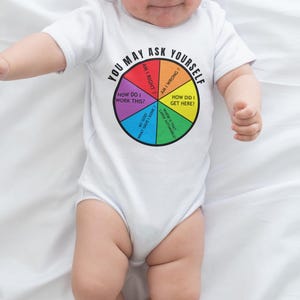 Puede incluir: Un body blanco para bebé con un diseño de diagrama circular colorido y el texto "You May Ask Yourself". El diagrama incluye preguntas como "How Did I Get Here?" y "Am I Right?". El bebé lleva una diadema blanca.