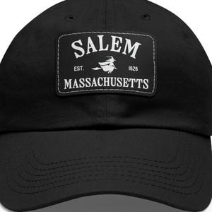 May include: Black baseball cap featuring a white patch with the word "Salem" above "Massachusetts". The patch includes a witch graphic and the text "Est. 1626". A classic, adjustable hat.