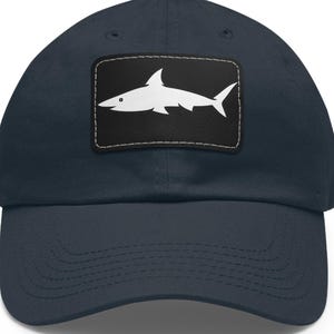 May include: Navy blue baseball cap featuring a white shark silhouette on a black rectangular patch. The cap has a curved brim and a structured crown. The shark design is a simple, clean graphic.