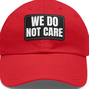 Könnte beinhalten: Eine leuchtend rote Baseballkappe mit gebogenem Schirm. Ein schwarzes rechteckiges Patch auf der Vorderseite zeigt den weißen Text "WE DO NOT CARE". Die Kappe besteht aus einem weichen Stoff und hat eine strukturierte Krone.