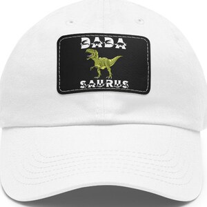 May include: White baseball cap with a black patch displaying the text "BABA SAURUS" and a green dinosaur illustration. The cap has a curved peak and a classic six-panel design.