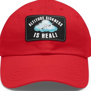 May include: A bright red baseball cap with a curved brim. The cap features a rectangular patch with the text "ALTITUDE SICKNESS IS REAL!" above an image of a plane in a cloud.