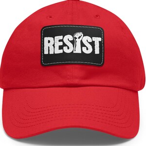 May include: Red baseball cap with a black patch that says "RESIST" in white letters with a raised fist graphic.