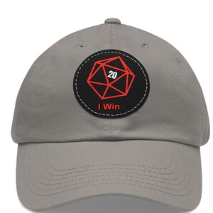 May include: A grey baseball cap with a curved peak. The cap has a round patch with a red 20-sided die design and the white text "I Win". The patch is on a black background.