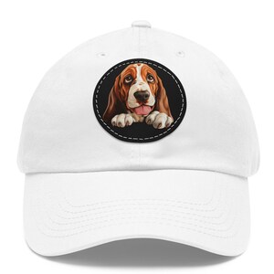 Könnte beinhalten: Eine weiße Baseballkappe mit einem runden Aufnäher, der eine Cartoon-Illustration eines Basset Hounds zeigt. Der Hund hat braunes und weißes Fell, und seine Zunge ist zu sehen. Der Aufnäher hat einen schwarzen Rand und weiße Nähte.