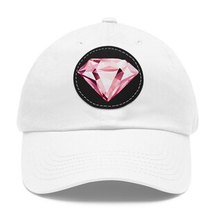 May include: White baseball cap with a pink diamond patch on the front. The diamond is surrounded by a black circle with a stitched border.
