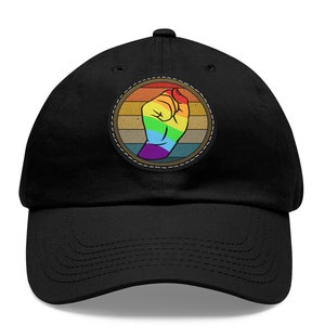 Op de afbeelding: Zwarte baseballpet met een cirkelvormige patch met een regenboogkleurige vuist tegen een gestreepte achtergrond. De patch is op de pet genaaid, die een gebogen klep heeft.