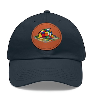 Op de afbeelding: Marineblauwe baseballpet met een ronde, bruine patch. De patch toont een kleurrijk, smeltend Rubik's Cube-ontwerp. De pet heeft een gebogen klep en een verstelbare sluiting.