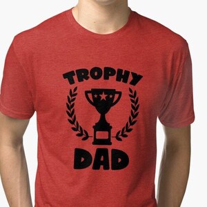 Può includere: T-shirt rossa con la scritta nera "TROPHY DAD", con una grafica di un trofeo all'interno di una corona d'alloro. La maglietta è girocollo e ha maniche corte.