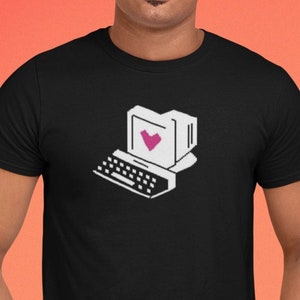 Puede incluir: Camiseta negra con un gráfico de ordenador retro y un corazón rosa en la pantalla. El diseño es en blanco y negro, creando un contraste con el color oscuro de la camiseta. La camiseta la lleva una persona.