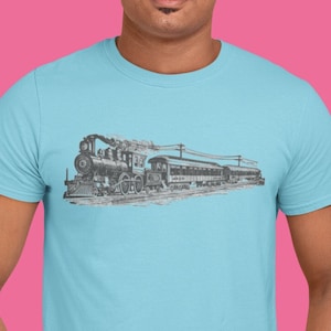 Puede incluir: Un hombre con una camiseta azul claro con una ilustración en blanco y negro de un tren de vapor en la parte delantera. Lleva pantalones vaqueros azules y tiene las manos en los bolsillos.
