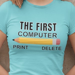 Puede incluir: Camiseta azul claro con el texto "THE FIRST COMPUTER" sobre un dibujo de un lápiz amarillo. Debajo del lápiz están las palabras "PRINT" y "DELETE". La modelo lleva un pantalón corto vaquero negro.