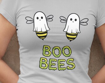 May include: A light gray short-sleeved t-shirt featuring a graphic of two bees wearing white ghost sheets. Below the bees, the text BOO BEES is printed in a lime green, dripping font. The shirt is paired with dark gray denim shorts and black tights.